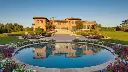 A luxurious brick house perched elegantly on a hill, featuring a sparkling swimming pool, a vibrant flower garden, and a spacious car park, all perfectly reflected in a serene body of water or a polished surface..webp